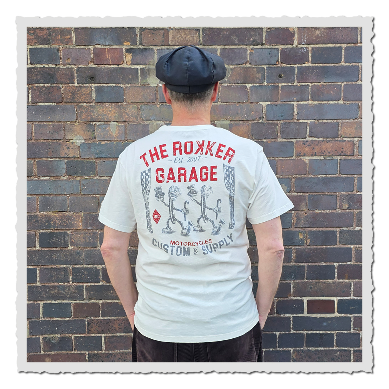 Mann von hinten in hellem T-Shirt mit rotem und grauem Rückenprint, vor mehrfarbiger Backsteinwand.